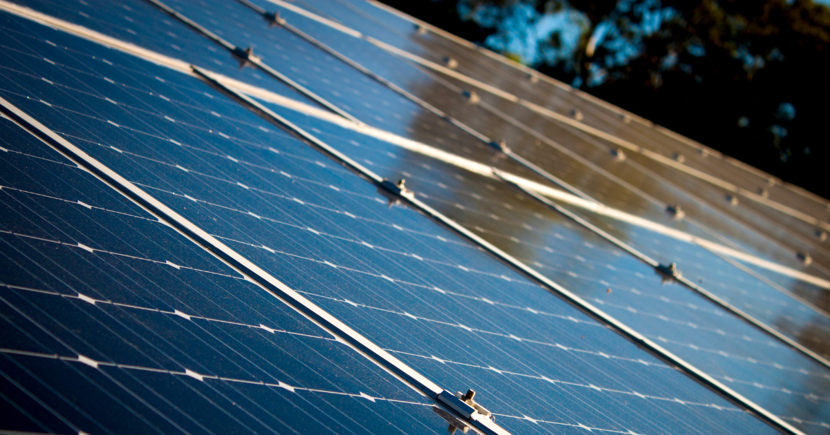 Vista lateral de módulos solares inclinados, refletindo luz em um ambiente ao ar livre com árvores ao fundo. A imagem reforça a sustentabilidade e o uso de energia solar.