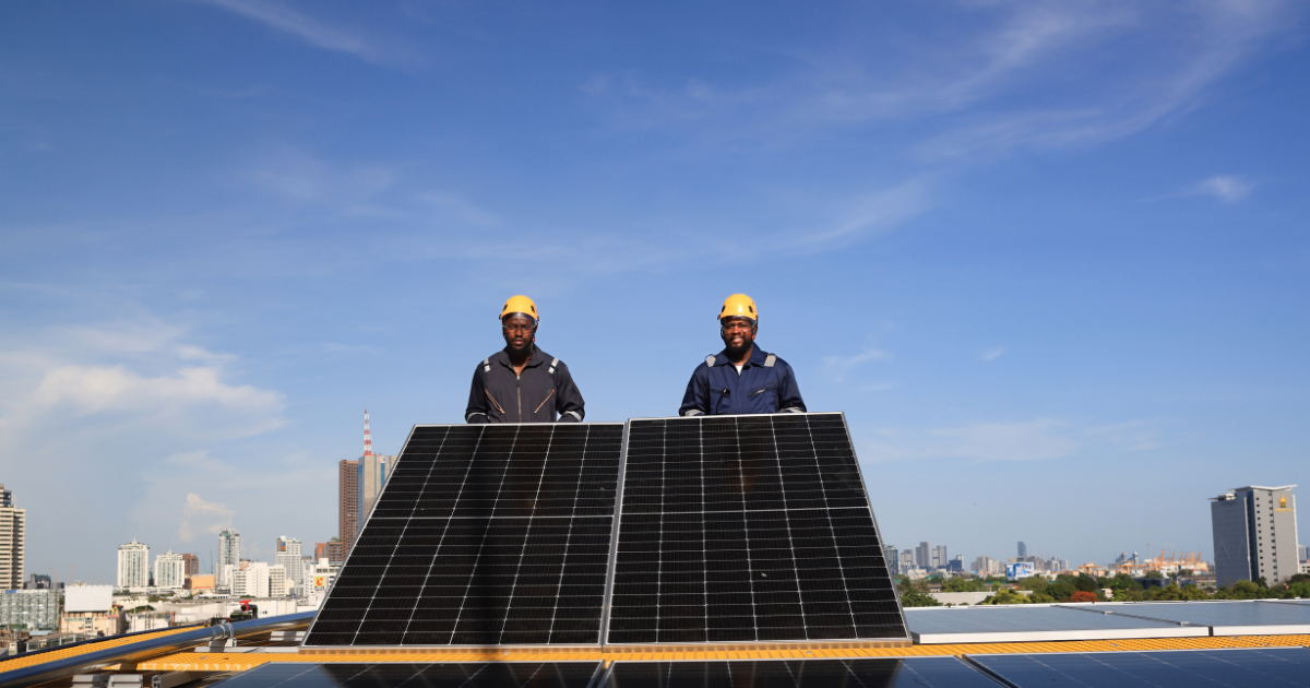  Dois profissionais com capacetes de segurança posam ao lado de painéis solares em um telhado, com a cidade ao fundo. 