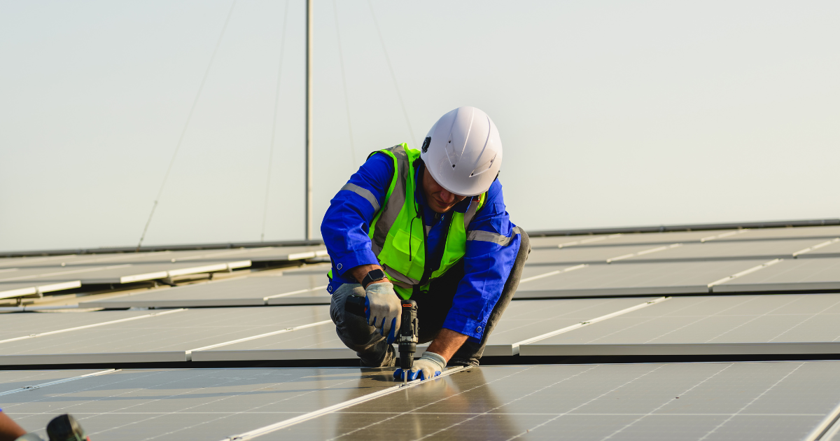 Integrador solar instala painéis solares em um telhado, usando capacete branco, colete refletivo e uniforme azul.