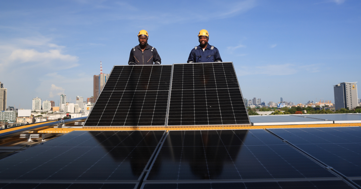 Dois integradores com capacetes amarelos estão em pé atrás de painéis solares em um telhado. Ao fundo, vê-se uma cidade com prédios e céu azul, destacando o uso de energia renovável.