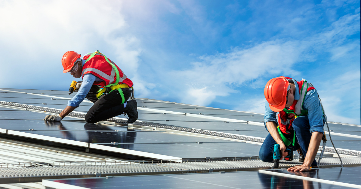 Dois integradores instalam painéis solares em um telhado, usando capacetes, coletes de alta visibilidade e equipamentos de proteção. O céu azul ao fundo destaca a energia limpa e renovável.