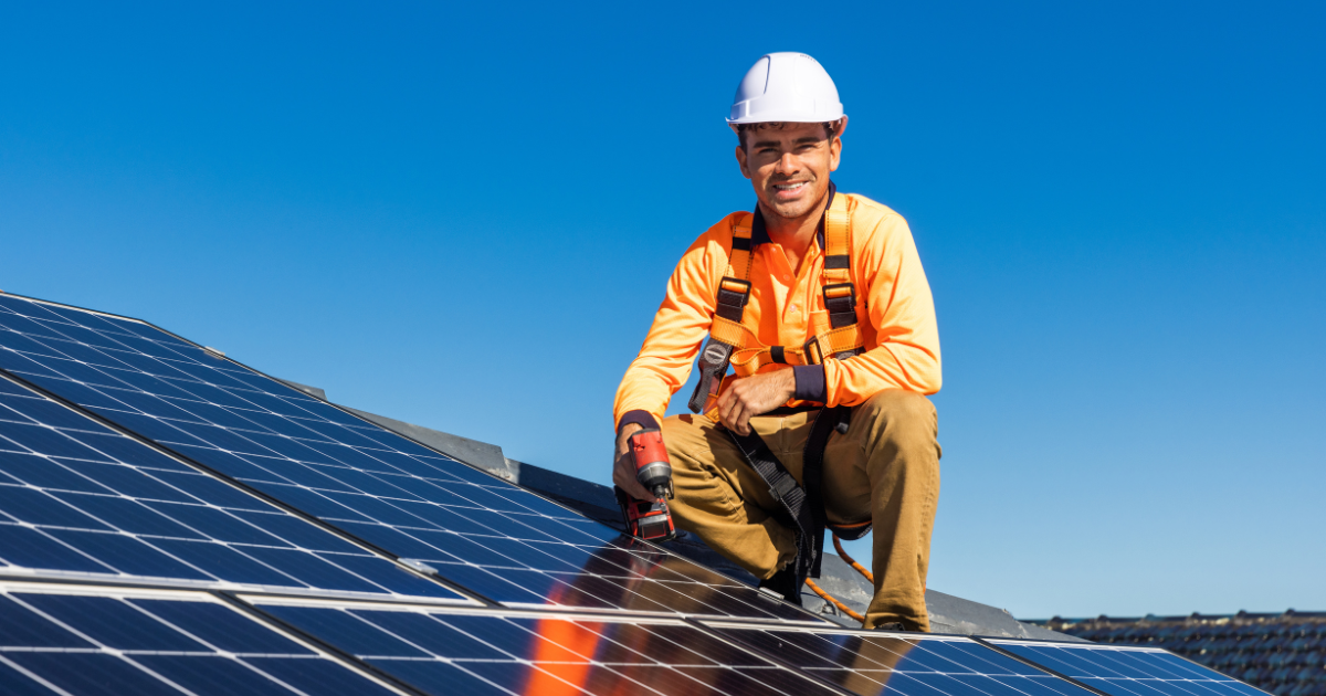 A imagem mostra um integrador solar sorridente instalando painéis solares, usando capacete, uniforme laranja e cinto de segurança.