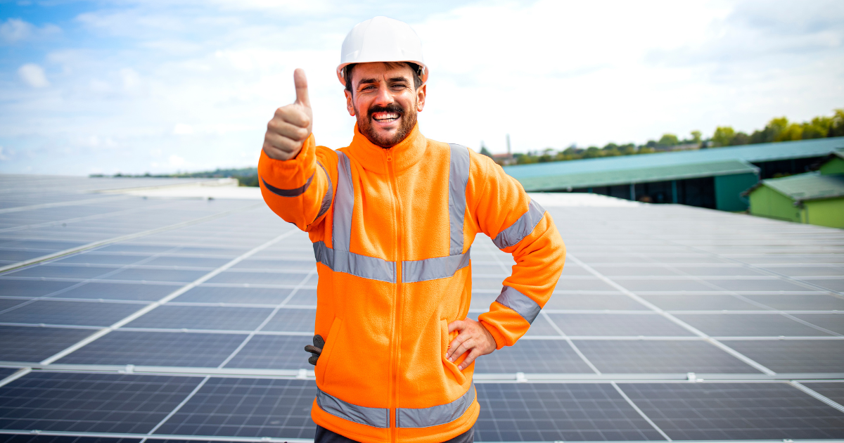 Integrador solar sorridente com capacete e jaqueta refletiva faz gesto de positivo (joinha) sobre um campo de painéis solares.