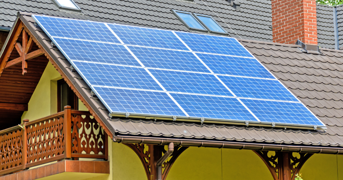 A imagem mostra uma casa com telhado inclinado coberto por painéis solares, destacando uma arquitetura com madeira trabalhada e uma chaminé de tijolos. 